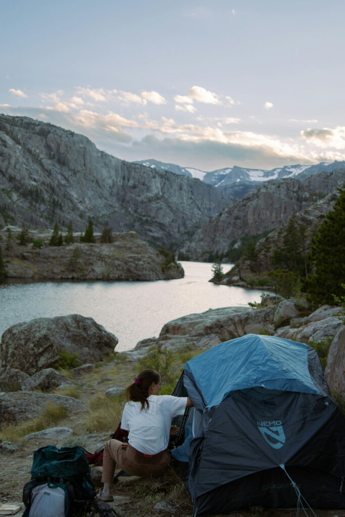 river leading to rocky mountains. camper sitting and reaching into a tent in the forground.